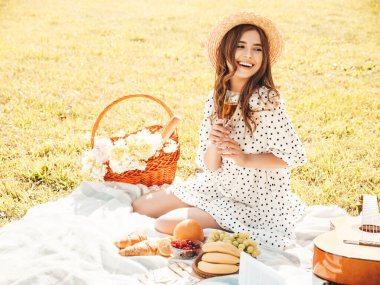 Moda elbisesi ve şapkası olan genç ve güzel hippi kadın. Kaygısız kadın dışarıda piknik yapıyor. Pozitif model çimlerin üzerinde oturuyor, meyve ve peynir yiyor, şampanya içiyor.