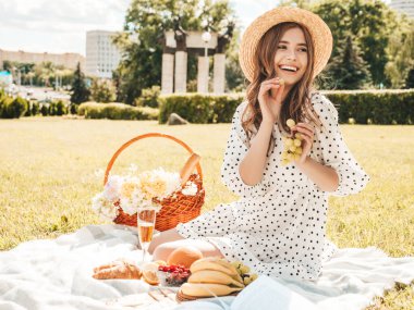 Moda elbisesi ve şapkası olan genç ve güzel hippi kadın. Kaygısız kadın dışarıda piknik yapıyor. Pozitif model çimlerin üzerinde oturuyor, meyve ve peynir yiyor, şampanya içiyor.