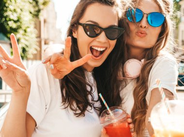 Moda kıyafetleri içinde gülümseyen iki güzel hippi kadın. Dışarıda poz veren kaygısız kadınlar. Pozitif modeller, plastik bardakta pipetle taze kokteyl içiyorlar ve içiyorlar.