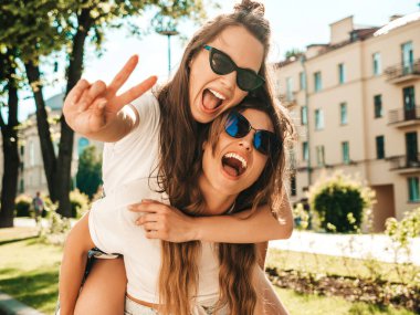 Son moda beyaz tişörtlü iki güzel, gülümseyen yeniyetme kadın sokak arka planında poz veren kaygısız kadınlar. Manken arkadaşının sırtına atlayıp, onu dışarı taşır.