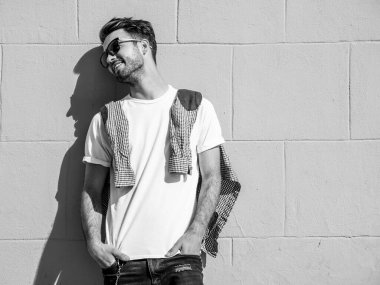 Yakışıklı, gülümseyen stil sahibi Lambersex modelinin portresi. Beyaz tişört giyen adam. Moda erkek, dışarıda güneş gözlüklü, duvarın yanında poz veriyor.