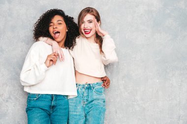 İki güzel, güzel, gülümseyen uluslararası hippi kadın, son moda yaz kot kıyafetleri içinde. Stüdyoda gri duvarın yanında poz veren kaygısız seksi kadınlar. Pozitif modeller eğleniyor. Arkadaşlık kavramı 