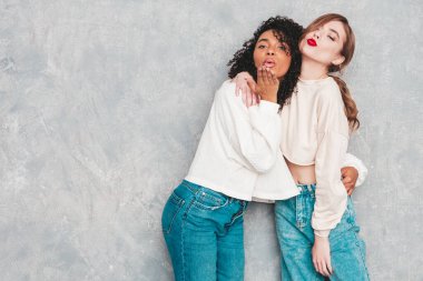 İki güzel, güzel, gülümseyen uluslararası hippi kadın, son moda yaz kot kıyafetleri içinde. Stüdyoda gri duvarın yanında poz veren kaygısız seksi kadınlar. Pozitif modeller eğleniyor. Arkadaşlık kavramı 