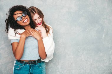 İki güzel, güzel, gülümseyen uluslararası hippi kadın, son moda yaz kot kıyafetleri içinde. Stüdyoda gri duvarın yanında poz veren kaygısız seksi kadınlar. Pozitif modeller eğleniyor. Arkadaşlık kavramı 