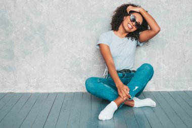 Saçları kıvırcık, güzel siyah bir kadın. Moda kotlar ve güneş gözlükleriyle gülümseyen bir model. Stüdyoda gri duvarın yanında oturan kaygısız seksi kadın.