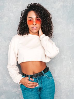 Saçları kıvırcık, güzel siyah bir kadın. Beyaz kotlu ve güneş gözlüklü, gülümseyen bir model. Stüdyoda gri duvarın yanında poz veren kaygısız seksi kadın. Bronzlaşmış ve neşeli 