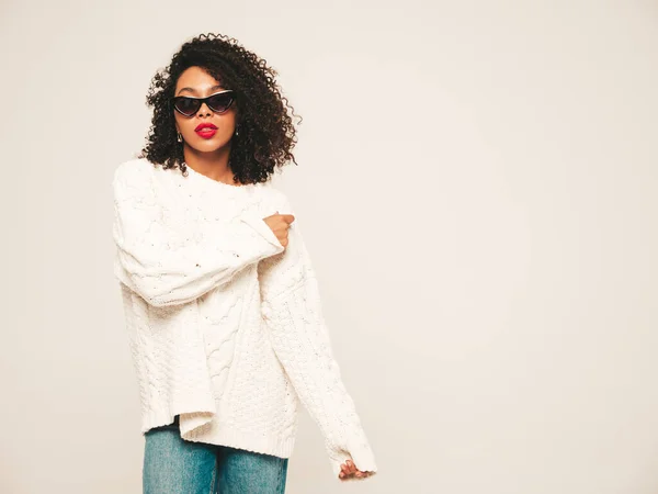 Beautiful black woman with afro curls hairstyle.Smiling model in white winter sweater and jeans clothes and sunglasses. Sexy carefree female posing on white background in studio. Tanned and cheerful