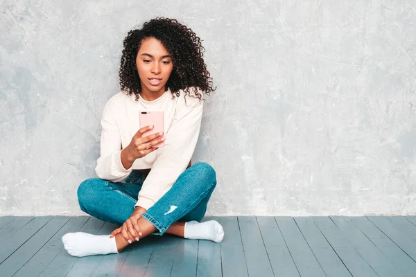 Beautiful black woman with afro curls hairstyle.Model in white trendy jeans clothes and sunglasses. Sexy carefree female sitting on the floor near gray wall in studio.Looking at cellphone. Using apps