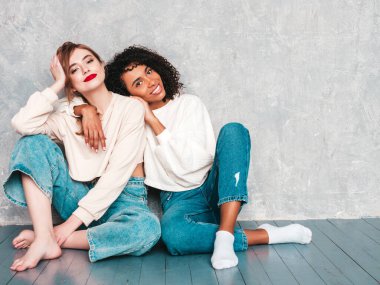 İki güzel, güzel, gülümseyen uluslararası hippi kadın, son moda yaz kot kıyafetleri içinde. Stüdyoda gri duvarın yanında oturan kaygısız seksi kadınlar. Pozitif modeller eğleniyor. Arkadaşlık kavramı 