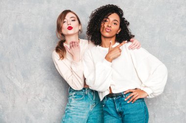 İki güzel, güzel, gülümseyen uluslararası hippi kadın, son moda yaz kot kıyafetleri içinde. Stüdyoda gri duvarın yanında poz veren kaygısız seksi kadınlar. Pozitif modeller eğleniyor. Arkadaşlık kavramı 