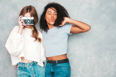 İki güzel, güzel, gülümseyen uluslararası hippi kadın, son moda yaz kot kıyafetleri içinde. Stüdyoda gri duvarın yanında poz veren kaygısız seksi kadınlar. Pozitif modeller eğleniyor. Retro kamerada fotoğraf çekmek