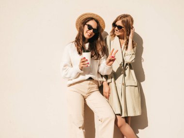 Son moda beyaz süveter ve palto giymiş iki güzel, gülümseyen hippi kadın. Sokakta şapkayla poz veren kaygısız seksi kadınlar. Pozitif modeller güneş gözlükleriyle eğleniyor.
