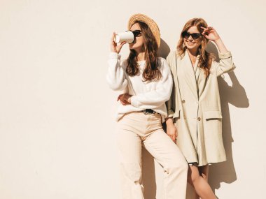 Son moda beyaz süveter ve palto giymiş iki güzel, gülümseyen hippi kadın. Sokakta şapkayla poz veren kaygısız seksi kadınlar. Pozitif modeller güneş gözlükleriyle eğleniyor.