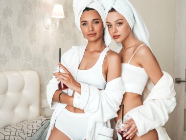 Beyaz bornozlu ve kafasında havlu olan iki güzel kadın. Otel odasında yatakta oturan kaygısız seksi mankenler. Taze kokteyl içecekler plastik bardakta samanla içecekler.