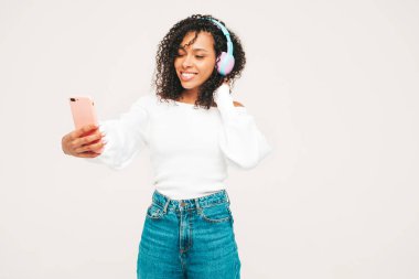 Saçları kıvırcık, saçları kıvırcık, süveter ve kot giyen, kablosuz kulaklıkla müzik dinleyen kaygısız bir kadın. Stüdyoda beyaz arka planda poz veriyor. Akıllı telefon tutuyor.