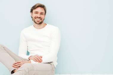 Beyaz süveter ve pantolon giyen yakışıklı, gülümseyen hippi, keresteci iş adamı modeli portresi. Moda tarzı bir adam stüdyoda açık mavi duvara karşı oturuyor.
