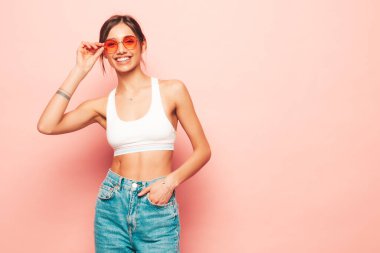 Beyaz formalı bluz ve kot pantolon giymiş güzel bir kadın. Seksi, kaygısız, neşeli manken içeride eğleniyor. Stüdyoda pembe duvarın yanında poz veren sevimli ve pozitif bir kadın. Güneş gözlüklü.