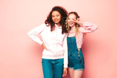 Moda kıyafetleri içinde iki güzel, gülümseyen uluslararası hippi kadın. Stüdyoda pembe duvarın yanında poz veren kaygısız seksi kadınlar. Pozitif modeller eğleniyor. Arkadaşlık kavramı 