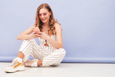 Yeni moda hippi kıyafetleri içinde gülümseyen güzel bir kadın. Stüdyoda mor duvarın yanında oturan kaygısız seksi kadın. Pozitif model içeride eğleniyor. Akıllı telefonu elinde tutarak. Uygulamalar kullanılıyor 