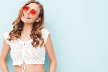 Modayı takip eden yaz hippi kıyafetleri içinde gülümseyen genç bir kadının portresi. Stüdyoda açık mavi duvarın yanında poz veren kaygısız bir kadın. Güneş gözlükleriyle evde eğlenen pozitif model. Neşeli ve mutlu.