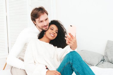 Gülümseyen güzel kadın ve yakışıklı erkek arkadaşı. Mutlu çok ırklı aile selfie çekiyor. Yatakta yatan ve beyaz iç mekanlarda sarılan çok ırklı modeller. Birbirinize sarılın.