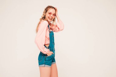 Genç ve güzel bir kadın kameraya bakıyor. Popüler bir bayan günlük kot pantolon içinde gülümsüyor. Stüdyoda gri arka planda poz veren pozitif ve komik bir model. Dilini güneş gözlükleriyle gösteriyor.