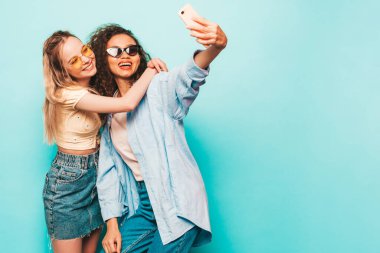 Moda kıyafetleri içinde iki güzel, gülümseyen uluslararası hippi kadın. Stüdyoda mavi duvarın yanında poz veren kaygısız seksi kadınlar. Pozitif modeller güneş gözlükleriyle eğleniyor. Selfie çekiyoruz.