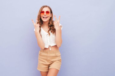 Son moda yaz hippi kıyafetleri içinde gülümseyen güzel bir kadının portresi. Stüdyoda mor duvarın yanında poz veren kaygısız seksi kadın. Pozitif model içeride eğleniyor. Güneş gözlükleriyle barış işareti gösterir