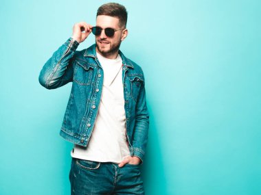 Yakışıklı, gülümseyen stil sahibi Lambersex modelinin portresi. Ceket ve kot giyen adam. Moda erkeği, stüdyoda güneş gözlüklü mavi duvarın yanında poz veriyor.