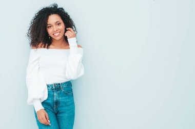 Saçları kıvırcık, güzel siyah bir kadın. Süveter ve kotlar içinde gülümseyen bir model. Stüdyoda açık mavi duvarın yanında poz veren kaygısız seksi kadın. Bronzlaşmış ve neşeli