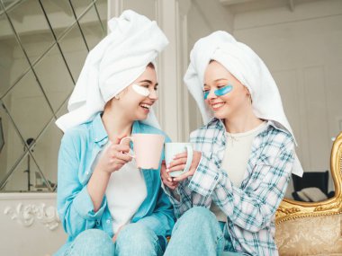 Gözlerinin altında bantlarla gülümseyen iki genç kadın. Lüks bir dairede yatağında oturan kaygısız mankenler. Evde havlularla kafalarının üstünde güzellik tedavisi yapıyorlar. Çay içiyor, bardak tutuyor.
