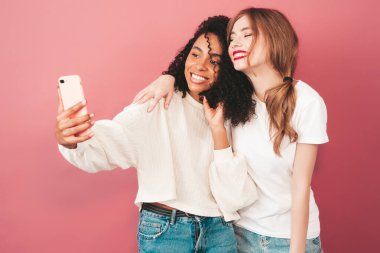 İki güzel, güzel, gülümseyen uluslararası hippi kadın, son moda yaz kot kıyafetleri içinde. Stüdyoda pembe duvarın yanında poz veren kaygısız seksi kadınlar. Pozitif modeller eğleniyor. Selfie çekiyoruz.