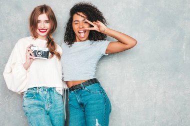 İki güzel, güzel, gülümseyen uluslararası hippi kadın, son moda yaz kot kıyafetleri içinde. Stüdyoda gri duvarın yanında poz veren kaygısız seksi kadınlar. Pozitif modeller eğleniyor. Retro kamerada fotoğraf çekmek