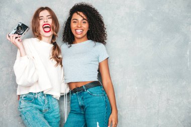 İki güzel, güzel, gülümseyen uluslararası hippi kadın, son moda yaz kot kıyafetleri içinde. Stüdyoda gri duvarın yanında poz veren kaygısız seksi kadınlar. Pozitif modeller eğleniyor. Retro kamerada fotoğraf çekmek