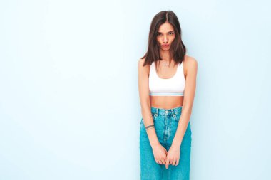 Beyaz formalı bluz ve kot pantolon giymiş güzel bir kadın. Seksi kaygısız model sabahının tadını çıkarıyor. Stüdyoda açık mavi duvarın yanında poz veren sevimli ve pozitif bir kadın. Ördek suratı yapar