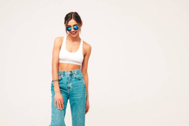 Beyaz formalı bluz ve kot pantolon giymiş güzel bir kadın. Seksi, kaygısız, neşeli manken içeride eğleniyor. Stüdyoda gri arka planda sevimli ve pozitif bir kadın poz veriyor. Güneş gözlüğü takıyor.