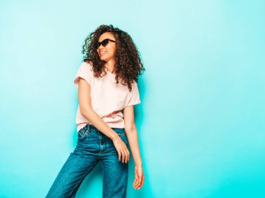 Saçları kıvırcık, beyaz tişörtlü ve kot pantolonlu güzel siyahi bir kadın. Stüdyoda güneş gözlükleriyle maviliğe yakın poz veren kaygısız seksi kadın. Bronzlaşmış ve neşeli 