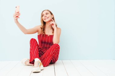 Yeni moda kırmızı takım elbiseli güzel, gülümseyen bir kadının portresi. Stüdyoda açık mavi duvarın yanında oturan kaygısız seksi kadın. Kapalı alanda eğlenen pozitif model. Selfie çekiyoruz.