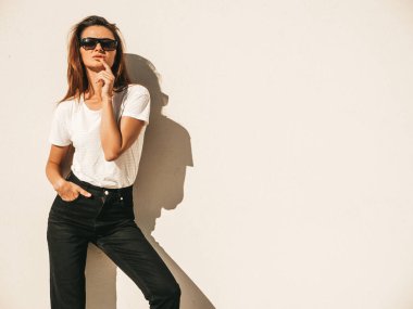 Güneş gözlüklü güzel gülümseyen modelin portresi. Yazlık hippi tişörtü ve kot pantolon giymiş bir kadın. Sokakta duvar kenarında poz veriyor. Komik ve pozitif kadın dışarıda eğleniyor.