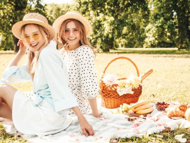 İki güzel, güzel, güler yüzlü yeniyetme yaz elbisesi ve şapkalı kadın. Piknik yapan kaygısız kadınlar. Çimlerin üzerinde oturan, şampanya içen, meyve ve peynir yiyen pozitif modeller.
