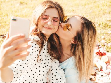 İki güzel hippi kadın, son moda yaz elbisesi ve şapkaları içinde. Kaygısız kadınlar dışarıda piknik yapıyor. Çimlerin üzerinde oturan, meyve ve peynir yiyen ve selfie çeken pozitif modeller.