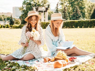 İki güzel, güzel, güler yüzlü yeniyetme yaz elbisesi ve şapkalı kadın. Piknik yapan kaygısız kadınlar. Çimlerin üzerinde oturan, şampanya içen, meyve ve peynir yiyen pozitif modeller.