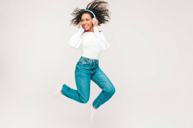 Kıvırcık saçlı, güzel siyah bir kadın. Süveter ve kot giyen, kablosuz kulaklıkla müzik dinleyen, kaygısız bir kadın. Beyaz arka planda stüdyoya atlamak. Dinamik hareket. 