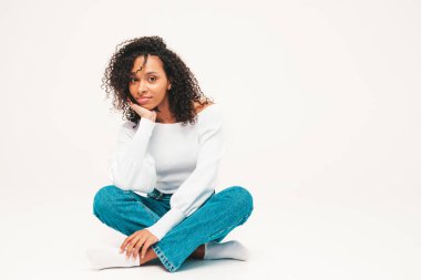 Saçları kıvırcık, güzel siyah bir kadın. Süveter ve kotlar içinde gülümseyen bir model. Stüdyoda beyaz arka planda oturan kaygısız seksi kadın. Bronzlaşmış ve neşeli 