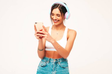 Kot pantolon giymiş, gülümseyen güzel bir kadın. Kablosuz kulaklıkla müzik dinleyen seksi tasasız model. Stüdyoda gri arka planda akıllı telefonunu kullanarak poz veren sevimli ve pozitif bir kadın.