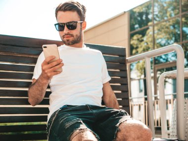 Yakışıklı, şık, havalı, lamberseksüel bir model. Beyaz tişört giymiş bir adam. Sokakta poz veren bir erkek. Cep telefonunda tutuyor, kaydırıyor ve mesaj atıyor. Akıllı telefondan mesaj yazar