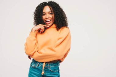 Afro kıvırcık saçlı, turuncu kapüşonlu ve kotlu güzel siyah kadın. Stüdyoda beyaz arka planda poz veren kaygısız seksi kadın. Bronzlaşmış ve neşeli