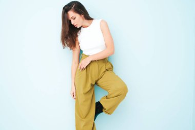 Moda kıyafetleri içinde gülümseyen genç ve güzel bir bayan. Stüdyoda açık mavi duvarın yanında poz veren kaygısız seksi kadın. Pozitif model içeride eğleniyor. Neşeli ve mutlu