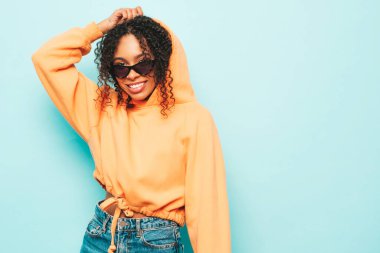 Afro kıvırcık saçlı, turuncu kapüşonlu ve kotlu güzel siyah kadın. Stüdyoda mavi duvarın yanında poz veren kaygısız seksi kadın. Güneş gözlükleriyle bronzlaşmış ve neşeli