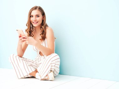 Yeni moda kırmızı takım elbiseli, gülen güzel bir bayan. Stüdyoda açık mavi duvarın yanında oturan kaygısız seksi kadın. Akıllı telefon ekranına bakan pozitif model. Uygulamalar kullanılıyor 